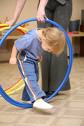 A picture of a young child ducking his head and walking through a hulahoop being held by an adult. 