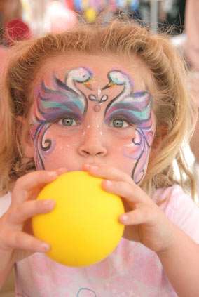 An image of a girl blowing up a yellow balloon. She has swans painted above her eyes with decorative designs painted down her cheeks. 