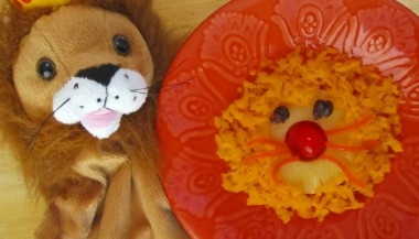 An image of a stuffed lion next to a bowl with a lion face made out of pineapple as the head, carrot as the hair, raisins as the eyes, cherry as the nose, and strings of carrot as the whiskers. 