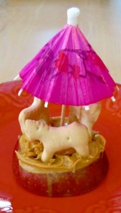 An image of an apple sliced horizontally with three animal crackers and an umbrella on top making it look like a carousel. 