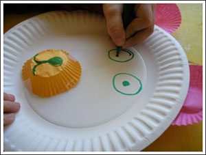 A picture of a child making a teddy bear face on a paper plate. Using green marker to make the eyes and a cupcake liner for the nose. 