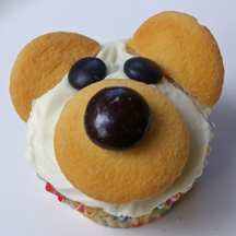 A picture of a cupcake that looks like a teddy bear. Vanilla waffers were used to make the nose and ears. White frosting on top. 