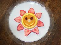 A picture of a pancake with a smiley face made out of fruit. There's cut up strawberries laid out around the pancake to represent the suns rays. 