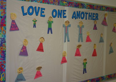 A bulletin board with images of different boys and girls on the board. It appears as if they were crafted out of construction paper and glued to the board. The heading of the bulletin board is "Love One Another." 