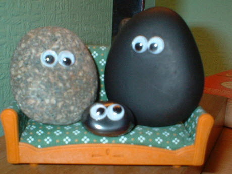 A picture of 3 rocks decorated with googly eyes sitting on a doll couch. 