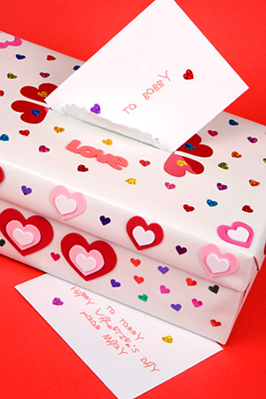 A shoe box decorated in small and medium colorful hearts. There is an opening to put valentines cards in and the picture shows a card being dropped into the opening. 