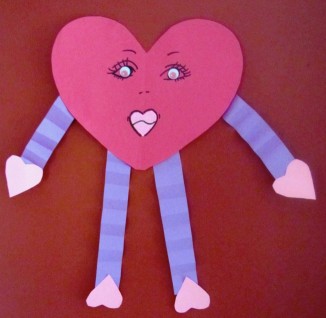 A heart cut from red construction paper with a feminine face on it. The arms are made from purple accordion construction paper and the hands and feet are made from pink heart construction paper cutouts. 