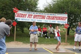 A picture of two people competing in a cherry pit spitting contest with people looking on. The sign in the background reads "Cherry Pit Spitting Championship."