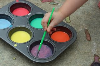 A picture of a muffin pan filled with different colored paint. A child's hand is dipping a paintbrush into the purple paint. 