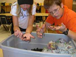 A picture of kids digging through a big tub of dirt and worms. The boy and girl are observing the worms they found in their hands. 