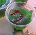 A picture of green Jell-o in a clear plastic cup with gummy worms in the Jell-o