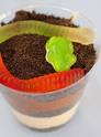 A worm themed snack idea that appears to be a worm on dirt. A picture of crushed Oreo cookies in a see through plastic glass with a gummy worm sitting on top. 