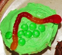 A cupcake with green frosting and green M&Ms with a red gummy worm on top. 