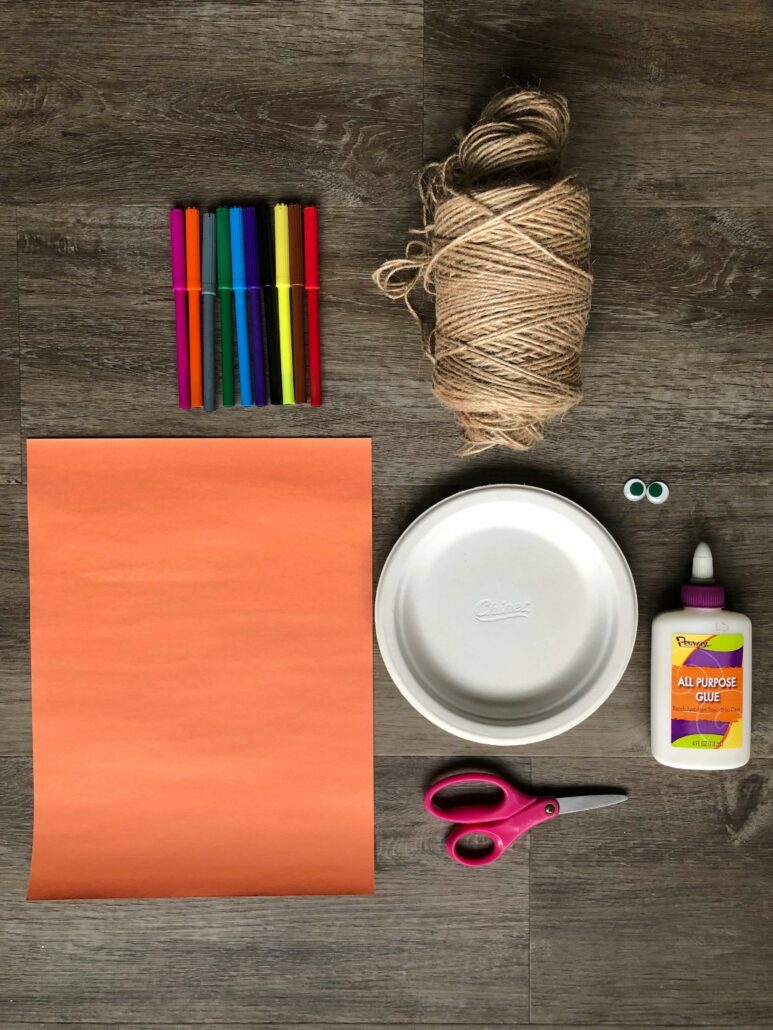 An image of all the materials used to make the paper plate face craft: markers, twine string, orange construction paper, paper plate, googly eyes, glue, and scissors. 