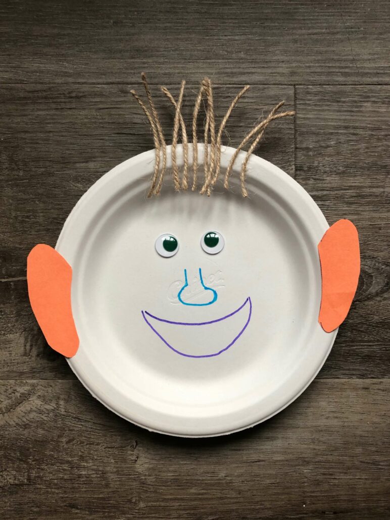 An image of the finished paper plate face craft. The paper plate has a purple mouth, blue nose, googly eyes, orange construction paper ears, and twine hair.